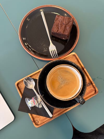 An overhead view captures a dessert and coffee arrangement on a teal-colored table in Le Havre, France. In the upper center, a round black plate with an orange-brown rim features a textured surface and holds a single rectangular chocolate brownie. A silver fork rests diagonally across the plate, tines pointing towards the upper left. Below this, a black ceramic coffee cup with a lighter interior, containing a dark brown liquid topped with light brown crema, sits on a matching black saucer. This coffee setup is positioned on a small, rectangular wooden tray with raised edges. Adjacent to the coffee cup on the tray, a silver spoon lies on a folded black napkin, next to a clear plastic-wrapped candy, identifiable as a "La Perruche" sugar cube. A portion of a white object, likely a laptop, is visible at the far left edge of the teal table. A subtle diagonal seam is present on the table's upper left surface, and a faint water mark or reflection is visible on the lower right of the table.