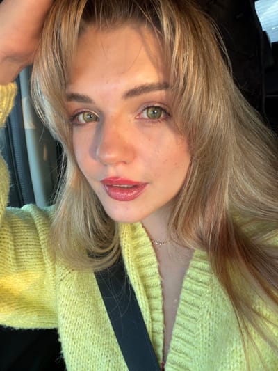 A close-up portrait features a young Caucasian female. She possesses long, light blonde hair styled with bangs, green eyes, and fair skin with scattered freckles visible on her nose and cheeks. Her lips are coated with a red-pink glossy product, and her eyelids exhibit a light, possibly gold-toned, cosmetic application. She is attired in a light yellow, possibly knitted, cardigan or sweater. A black seatbelt strap is positioned diagonally across her chest. A delicate necklace with small letter beads spelling "princess" rests on her neck. Her left hand is partially visible in the upper left quadrant, appearing to touch her hair. The subject looks directly at the camera. The background is dark, out of focus, and indistinct, characteristic of an interior or vehicle setting. A faint vertical light-colored element is present on the far left. The lighting is natural, casting subtle shadows. The exact location within the city of Азьмушкино, Russia, cannot be determined from the visual information in the image.