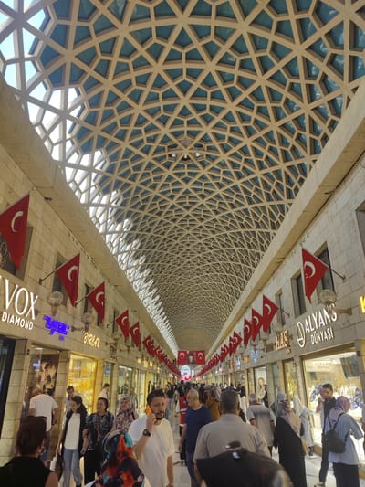 This image depicts a bustling indoor marketplace, likely in Osmangazi, Turkey, characterized by its distinctive geometric ceiling architecture. The ceiling features an intricate lattice of beige structural elements interspersed with light blue panels, creating a diamond-like pattern overhead. Along the sides of the walkway, shops display their goods behind glass storefronts. Prominently displayed on the building facades are numerous Turkish flags, featuring a red background with a white crescent moon and star.

The marketplace is populated with a diverse crowd of people walking in both directions. Many individuals are dressed in casual attire, while some women are wearing headscarves. Several people are looking into shop windows, while others are engaged in conversation or walking purposefully. One man in the foreground is holding a mobile phone to his ear. Shop signs visible include "VOX DIAMOND," "ZUHAL," "BIRLHAN," and "ALYANS DÜNYASI," suggesting a concentration of jewelry or luxury goods stores. The overall atmosphere appears to be lively and active, with natural light filtering through the patterned ceiling.