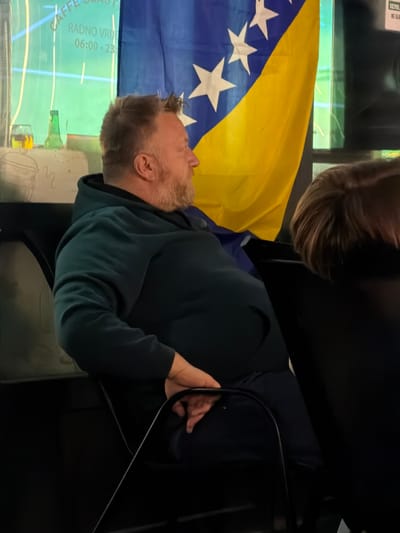 In Nević Polje, Bosnia and Herzegovina, a middle-aged man with a beard is seated in a chair, facing right. He is wearing a dark teal hooded sweatshirt and dark pants. His arms are resting on his lap, with his left hand partially visible. To his right, a Bosnian flag is displayed prominently. In the background, a window shows a green-tinted advertisement for "CAFFE SLAS." with operating hours "RADNO VRIJEME 06:00-23:00." A glass with a yellow liquid and a green bottle are also visible on a surface behind the man. The scene appears to be indoors, possibly in a cafe or public establishment, with other patrons hinted at by the presence of another person's head and shoulders to the right of the frame.