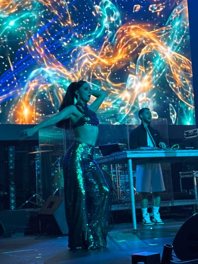 In Misaktsieli, Georgia, on a stage bathed in vibrant blue and green lighting, a woman with dark hair pulled into a ponytail performs. She wears a sparkling, iridescent green halter top and matching, floor-length sequined skirt. Her arms are outstretched, and her body is positioned in a dynamic dance pose. Behind her, a large screen displays a psychedelic, abstract pattern of swirling blue, teal, and orange light. To the right, a man stands behind a DJ console, wearing a dark jacket over a white shirt and light-colored shorts. He appears to be engaged with the equipment, possibly mixing music. The stage also features various musical instruments and speakers, suggesting a live performance. The overall atmosphere is energetic and visually stimulating, characteristic of a concert or club event.