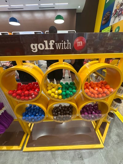 The image depicts a display of M&M candies in cylindrical bins of various colors at the M&M's store in Lake Buena Vista, Florida, United States. The candies are arranged by color, including orange, purple, green, yellow, brown, red, and blue. A sign adjacent to the display reads "golf with m" and has text in Spanish, reading "unwrap the yum!". Other signs with the M&M's logo are visible.