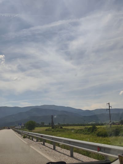 The image captures a daytime scene from a vehicle traveling on a road, offering a panoramic view of a mountainous landscape. In the foreground, the asphalt road extends into the distance, bordered on the right by a continuous metal guardrail. Beyond the guardrail, a vibrant expanse of green fields and foliage stretches towards a range of majestic mountains that form the backdrop, appearing hazy in the distance. The sky above is partly cloudy, featuring wispy white and gray clouds against a light blue backdrop.

Several objects populate the scene: utility poles and power lines are visible on the right side, cutting across the mid-ground. On the left side of the road, a blue rectangular sign is prominent, displaying symbols for "P" (parking) and "WC" (restrooms) with the text "300 M" beneath, indicating facilities ahead. Another sign below it shows "Kayatur". In the distance, other vehicles can be seen on the road, suggesting ongoing travel. The setting is likely a rural or semi-rural area, consistent with the provided location context of Skravena, Bulgaria. The weather appears fair and clear, suitable for a journey through this scenic environment.