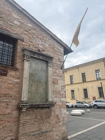 An urban street scene in Foligno, Italy, depicts a stone and brick building on the left, featuring a large, ornate rectangular frame enclosing a weathered religious fresco or mural, covered by a reflective pane. The artwork depicts multiple figures, including a central Madonna and child. Above the fresco, a grated window and a small, partially legible brown plaque are visible. A faded yellow, pointed banner extends diagonally from the building's upper corner on a black pole. The foreground is a paved area with dark gray pavers, delineated by white lines, containing white elongated concrete bollards and spherical concrete bollards. Several modern vehicles, including a white Fiat 500, a silver hatchback, and a white SUV, are parked on the right side of the paved area. In the background, a three-story building with a pale yellow stucco facade and multiple dark-framed windows stands under an overcast sky. A yellow streetlamp is affixed to the background building. No people are present.