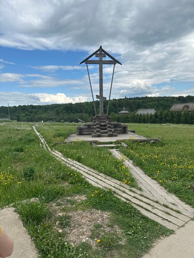 A tall wooden structure resembling a cross and a well stand in a grassy field in Vzglyadnevo, Russia. The sky is partly cloudy. A wooden pathway with slats runs through the field. A forest and several buildings are visible in the background. A paved road appears in the foreground. The grass is green and interspersed with yellow flowers.