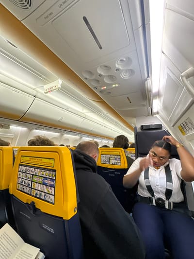 The image displays the interior of a Ryanair airplane cabin, captured from an aisle seat looking towards the front or middle of the aircraft. The cabin is brightly lit by internal lights, suggesting it is either daytime or that the cabin lights are fully on.
In the foreground, a cabin crew member is visible, wearing a white uniform shirt with epaulets, a harness, glasses, and a gold watch. They appear to be adjusting their hair or attire while seated. Behind them, several passengers are seated in the characteristic yellow and dark blue Ryanair seats. One passenger in the nearest row is seen from behind, wearing headphones, while others are partially visible further down the aisle. Each seatback features a safety information card with various pictograms illustrating emergency procedures.
Above the seating area, overhead bins line the cabin, many displaying the "RYANAIR" logo. An illuminated "← EXIT →" sign is prominent, indicating the location of an emergency exit. Overhead panels include individual air vents, reading lights, and call buttons.
Visible text includes "RYANAIR" in several locations, "← EXIT →," and "IMPORTANT NOTICE" on the safety card. Other smaller texts discernable from the OCR are "Life Vest," "Push Here," and the number "28" on overhead panels. The safety card also contains fragments like "Legal Requirement," "IN PANEL," "CUR HEAD," and "NOVE YOU."
The scene depicts a typical commercial flight environment, with passengers and cabin crew present. The provided context indicates that this flight is associated with Weeze, Germany, likely either departing from or arriving at Weeze Airport.