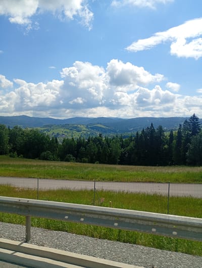 The image depicts a scenic view of the outskirts of Rdzawka, Poland. In the foreground, a metal guardrail with a grey metallic finish is visible, positioned along a roadside with a gravelly surface. Beyond the guardrail, a strip of green grass transitions into a road with a wire fence lining its edge.
Further into the scene, a dense forest of evergreen and deciduous trees stretches across the middle ground. Beyond the forest, rolling hills covered in lush green vegetation create a picturesque backdrop. Small buildings, possibly houses and other structures, are scattered across the slopes of these hills, suggesting a rural or semi-rural settlement. The sky is a vibrant blue, adorned with fluffy white cumulus clouds, indicating a bright and pleasant day. The overall impression is one of natural beauty and tranquility.