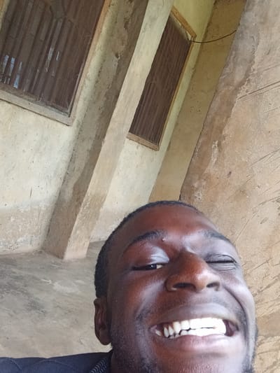 A close-up, low-angle selfie shows a Black male with dark skin and short black hair, grinning widely with his eyes squinted. He has a dark shirt on. The background is an outdoor, weathered concrete structure with two windows. The windows have wooden frames and metal bars, with the glass obscured. The concrete walls and pillars appear aged and rough. The scene is likely in Mbar, Nigeria, given the architectural style and building materials common in the region.
