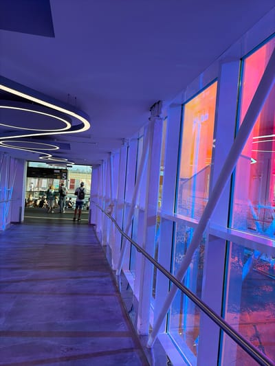 The media depicts an indoor hallway illuminated with purple and blue lights, with a glass ceiling that allows colored light to filter through. The hallway appears to be a section of an airport terminal, suggested by the presence of people queuing near a gate sign.

Subjects include several individuals, some of whom are waiting in line or walking. One person is standing, looking towards the gate. Another group is seated or crouched near the gate. An abstract white light sculpture is mounted on the left wall. The scene is characterized by modern architecture, with white beams supporting the glass ceiling and a polished floor.

The activity is people waiting for a flight. The setting is an airport terminal, possibly at Lamezia Terme, Italy, given the location context. The lighting suggests it might be evening or a deliberately stylized environment. The weather is not discernible from the interior shot. There is no discernible emotion conveyed by the people.

Visible text includes "BRUXELLES" and "Gate 5" on a digital display board, along with flight numbers and times, confirming the airport setting. There are also inscriptions on the light sculpture, but they are not legible.