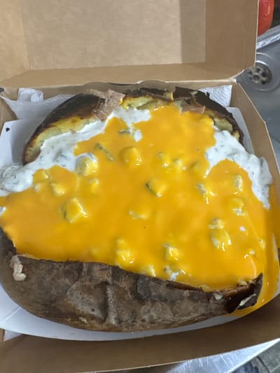A close-up, overhead shot captures a generously topped baked potato served in a brown cardboard take-out box. The potato is split open, revealing its fluffy interior, and covered with a thick layer of bright orange-yellow melted cheese sauce. Mixed within the cheese sauce are numerous small, lighter yellow or white chunks, possibly pieces of cheese or another ingredient. A contrasting white, creamy sauce, appearing similar to sour cream or ranch dressing with visible green specks (likely herbs), is also spread over the potato, particularly on the left side, partially covered by the cheese sauce.
The potato skin is dark brown and appears crispy from baking. A white paper napkin is tucked underneath the potato within the box. The cardboard box itself is plain brown, suggesting a casual meal or take-away service. In the blurred background, a metallic kitchen sink with a circular drain is visible, along with a red, rounded object, possibly a tomato. The setting appears to be indoors, likely a kitchen counter. No specific activity is depicted, but the prepared food suggests it is ready for consumption. There is no visible text in the image. The photo was taken in Нареченски бани, Bulgaria.