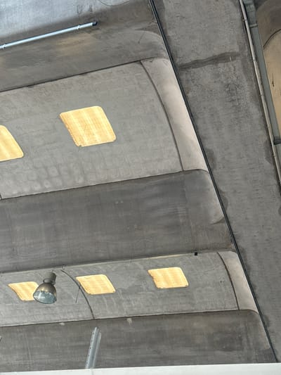 This is an eye-level, medium shot of the underside of a concrete overpass structure in Fontevivo, Italy. The concrete is weathered, with visible lines and imperfections, and is illuminated by several square, recessed lights that emit a warm, yellow glow. A metal light fixture, possibly a high-bay lamp, hangs from the ceiling. The perspective is looking upwards, emphasizing the architectural elements of the overpass. The scene is devoid of any people, animals, or vegetation, focusing solely on the man-made structure and its lighting.