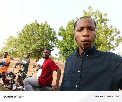 The image shows five men in an outdoor setting near Gidan Kogi, Nigeria. One man is taking a selfie, holding the camera at arm's length and obscuring part of the scene. He has a small stick or piece of straw in his mouth and wears a dark button-down shirt. Behind him, four other men are visible, some near motorcycles. One man is seated on a motorcycle wearing a red shirt and grey pants. Another man stands beside him wearing a white shirt. To the left, one man wears an orange t-shirt. Further left, another man in a green shirt is standing at the front of a motorcycle. The environment includes trees and what appears to be a dry, dusty ground.