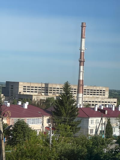 The image shows a panoramic view of Bely Klyuch, Russia. In the background, a tall industrial chimney with red and white stripes stands against a clear blue sky. To the left of the chimney is a large, multi-story concrete building with numerous windows. In the foreground, several residential buildings with red roofs and white chimneys are visible, surrounded by lush green trees. Electrical wires are strung across the scene, and a bird can be seen flying in the sky near the chimney. The overall impression is of an urban landscape with a mix of industrial and residential structures under a bright sky.