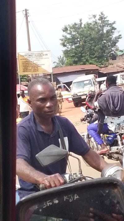 The image depicts a daytime street scene in Igbo Ukwu, Nigeria, characterized by local commerce and transit. In the immediate foreground, an adult male with short dark hair and a dark blue polo shirt operates a motorcycle, his hands gripping the handlebars. A black motorcycle tank/dashboard with "356 KJA" inscribed on it is partially visible. His gaze is directed slightly off-camera to the right.

In the midground, a white rectangular sign, supported by a wooden utility pole, advertises "ST. JOHN'S COMMUNITY CHILDREN WELFARE HOME (ORPHANAGE)," listing categories like "ORPHANS," "HOMELESS RANGES," "LONELY ADULTS," "VULNERABLE ADULTS," and "DEPRESSED ADULTS." To the right of the sign, a male figure in an orange and white shirt is partially visible, and further back, another individual in camouflage uniform walks. A white passenger van is parked behind them. To the right of the main subject, another motorcycle carries two male riders; the passenger wears a dark grey hooded top over patterned light-colored shorts/trousers and red flip-flops.

The background features simple, single-story buildings with reddish-brown corrugated iron roofs, indicative of local architecture. Lush green trees and vegetation are visible against a bright, hazy sky. The ground appears to be unpaved, suggesting a dusty road or market area.