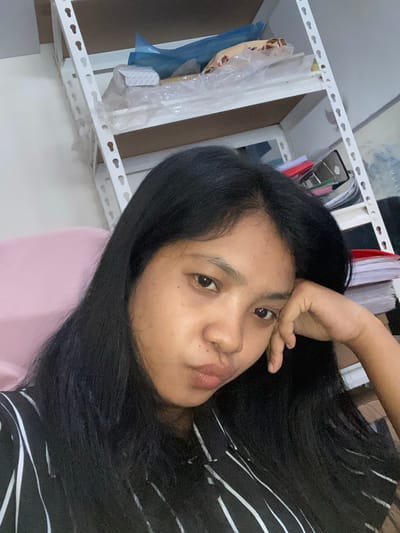 A medium, close-up shot depicts a woman with dark, straight hair. She is looking directly at the camera with a somewhat pensive expression. Her hand rests on her forehead. She is wearing a black and white striped shirt. Behind her, a white metal shelf is visible, containing various items wrapped in plastic. A pink surface is also visible behind her. The location is speculated to be in the city of Negara, Indonesia.