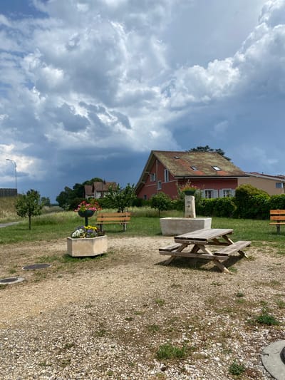The image depicts a public space in Boudry, Switzerland. In the foreground, a gravel path runs along the right side of the image. Along the path are visible two circular manhole covers and a concrete flower planter containing mixed flowers (white, yellow, and purple). Further along the path, a wooden bench is situated near a concrete water fountain. Behind the fountain, a red and tan building is visible. The background is dominated by a cloudy sky with varying shades of gray and white, suggesting possible stormy weather. A lamppost is visible on the left side of the image. There are no people visible in the image.