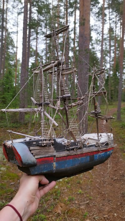 In a forest setting with tall pine trees and mossy ground, a person's hand is holding a weathered model of a sailing ship. The model depicts a historical vessel with multiple masts, rigging, and a hull painted in dark blue and red stripes, with some areas of decay and exposed wood. The ship's details, like the miniature sails and deck structures, suggest a past era of maritime travel. The person's hand, with a maroon bracelet on the wrist, is positioned to support the model from underneath, showcasing it against the natural backdrop. The lighting suggests it is daytime, possibly overcast, given the soft shadows. The overall mood is serene, with the intricate model contrasted against the wild, natural environment. No visible text is present in the image.