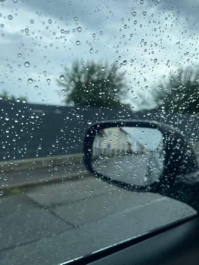 This is a photograph taken from inside a car looking through a rain-streaked window. Water droplets of varying sizes are scattered across the glass, obscuring the view of the outside. In the mid-ground, a dark-colored car with a side-view mirror is visible. The mirror reflects a blurry image of houses and a street. Beyond the car, trees and a building are visible through the rain. The sky appears overcast and cloudy. The photograph is likely taken in Geiselhöring, Germany, given the context, but no specific landmarks or identifying features in the image confirm this location.