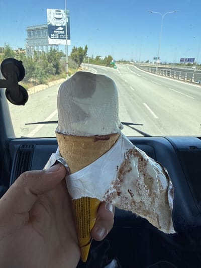 The image, rotated 90 degrees counter-clockwise, depicts a first-person view from inside a vehicle, likely a car or truck. A light-skinned hand is prominent in the lower right, holding a melting ice cream cone. The ice cream appears to be a generous scoop of a light-colored, possibly vanilla or caramel flavor, streaked with brown, and is actively dripping down the cone. The wrapper, a combination of yellow and white, is peeled back, revealing a crispy cone and fine print in multiple languages, which is largely illegible.

Through the vehicle's windshield, which shows some specks, a bright outdoor scene is visible. A wide, multi-lane highway stretches horizontally across the background, featuring clear white lane markings. The sky above is a brilliant, cloudless blue. Along the roadside, numerous green trees and bushes indicate lush, possibly Mediterranean, vegetation. Distinctive lampposts with "Y"-shaped tops are spaced along the road. In the mid-ground on the left, a large vertical billboard is visible, partially obscured by reflections. The billboard prominently displays "BOAT SEA" and the dates "08-10 May 2025," alongside smaller, unreadable text.

The interior of the vehicle shows a dark dashboard or door panel in the lower center and right, and a dark, suction-cup mounted device in the lower left. The scene is brightly illuminated, indicating a clear, sunny day. The melting ice cream suggests warm or hot weather. This moment of refreshment during travel is set in Aradippou, Cyprus, aligning with the sunny conditions and landscape.