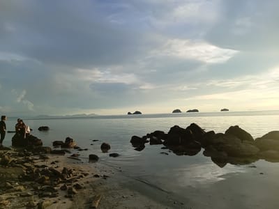 In Baan Pang Ka, Thailand, a tranquil beach scene unfolds as the sun begins to set, casting a soft golden light across the calm sea. The foreground is dominated by a rocky shoreline, with dark, wet stones scattered across the sandy beach and extending into the shallow water. Several dark rocks are partially submerged, their reflections shimmering on the water's surface. In the mid-ground, two figures are seated on a larger rock near the water's edge, one appearing to embrace the other. A third person stands to the left of the seated figures, facing the water. In the distance, several small, dark islands rise from the horizon, silhouetted against the pale, cloud-streaked sky. The overall atmosphere is peaceful and serene, suggesting a moment of quiet contemplation by the sea.
