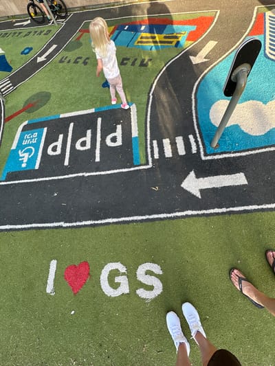 The image depicts an outdoor children's play area with a painted asphalt surface designed to resemble roads and buildings. In the center, a young blonde girl in a white t-shirt and light pink pants stands facing away from the camera, looking at a road design. The road features white lines, arrows, and symbols. To the left of the girl, a sign with Hebrew text and a wheelchair accessibility symbol is visible. Further down, a series of white letters "p" are painted on a black surface, indicating parking spaces. In the lower portion of the image, the words "I❤GS" are painted in white and red on a green artificial turf surface. Two pairs of feet are visible: one wearing white sneakers, and another wearing black flip-flops. The overall scene suggests a public playground in Givat Shmuel, Israel, as indicated by the letters "GS" in the painted text.