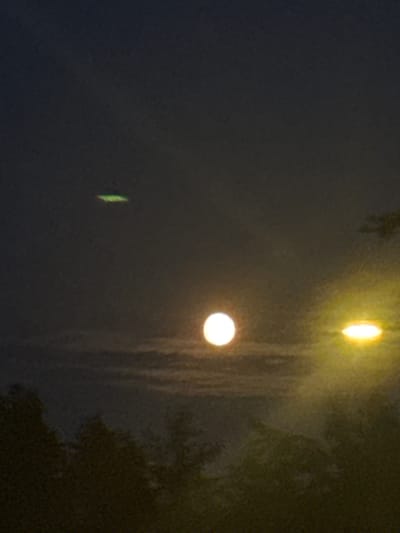 This is a nighttime photograph taken in Neumünster, Germany, depicting a sky with celestial bodies and artificial lights. The main focus is on a bright, circular celestial object, possibly the moon, positioned in the mid-ground. To its right, there is a bright, oval-shaped artificial light source. Further up and to the left of the main celestial object, a green, elongated light is visible, possibly an aircraft or drone. The foreground features a silhouette of trees and foliage against the dimly lit sky. The overall impression is that of a night sky with visible atmospheric conditions and man-made light pollution.