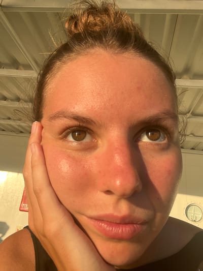A close-up, low-angle shot shows the face of a young woman with her right hand resting on her cheek. Her hair is pulled up into a messy bun, with strands falling around her face. She is looking upwards and slightly to her right. The setting appears to be outdoors, possibly on a boat or under a structure with a ribbed ceiling visible at the top of the frame. Sunlight casts a warm glow on her skin, highlighting a flush on her cheeks. In the background, a circular window with internal panes is visible on a white structure, and a red fire extinguisher sign is partially visible on the left. The image is taken in Batas Desa Skotong Timur, Indonesia.