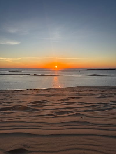This image depicts a serene sunset over the Dune of Pilat, located in La Teste-de-Buch, France. The foreground is dominated by the undulating, rippled sand dunes, casting long shadows that emphasize their textured surface. The middle ground features a body of calm water, likely the Arcachon Bay, reflecting the warm hues of the setting sun. A narrow strip of land or sandbar is visible in the bay, with some vegetation or structures. In the distance, the horizon is marked by a faint line of land. The sky transitions from a pale blue at the top to vibrant shades of orange and yellow near the horizon, where the sun, a bright orange orb, is setting. A few wispy clouds are scattered across the upper sky, and a small sailboat can be seen on the water. The overall scene is tranquil and picturesque, showcasing the natural beauty of this popular tourist destination.