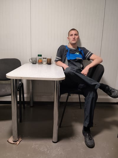 A young man, appearing to be in his early twenties, is seated at a white, speckled table with chrome legs. He is wearing dark blue overalls over a grey t-shirt with a green emblem on the chest. His legs are crossed, and his right arm rests on his lap. A dark grey chair is positioned to his left, and a black chair is partially visible behind him. On the table, there are three small containers, one of which appears to be a jar of spices, and two mugs, one clear and one brown, each with a spoon. The background is a white, corrugated wall. The floor is concrete and appears somewhat worn. The overall environment suggests a break room or a simple dining area, possibly within an industrial or workshop setting. The city of Шклоў, Belarus, is not directly identifiable from the visual cues within the image itself, and therefore cannot be determined as the location.