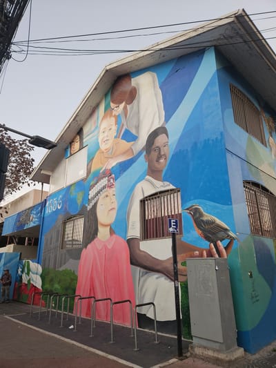 The image shows a vibrant mural painted on the side of a building in San Fernando, Chile. The mural depicts several people, including children and adults, in various poses and styles. One part of the mural features a young girl wearing a traditional indigenous headdress, a world map, and the year "1856". Another section shows a young boy and a woman, possibly a parent or teacher, interacting with him. A smiling young man is also prominently featured, as is a bird perched on a hand. The building itself appears to be a multi-story structure with windows that have metal bars. In the foreground, there is a bike rack and a utility box. The overall scene suggests a community art project or a cultural representation.
