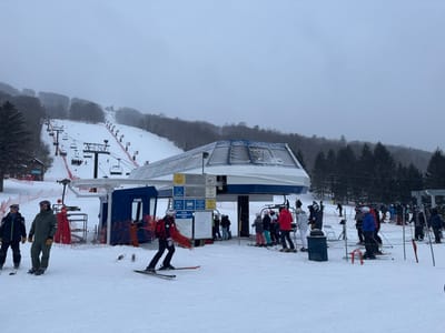 The image depicts a busy winter scene at the base area of Bristol Mountain Ski Resort in Canandaigua, New York, United States. In the foreground, a blue and white enclosed high-speed detachable chairlift terminal stands centrally, with snow covering its roof and various informational signs affixed to its structure. To the left of the terminal, two adult males, dressed in winter ski attire, stand on skis. To the immediate right of the terminal, an individual identified as ski patrol, wearing a red jacket and black pants, moves on skis while carrying a coiled red rescue sled. Further to the right, a queue of multiple skiers and snowboarders, including adults and children in full winter gear, waits for lift access, guided by red netting barriers. A dark blue waste bin is positioned near the queue. In the midground, multiple chairlift lines, with towers and occupied chairs, ascend a snow-covered mountain slope. Red safety netting delineates sections of the ski trails. The background reveals densely forested mountain slopes, primarily with evergreen trees. The sky is uniformly overcast and grey, indicating a cloudy or snowy day.