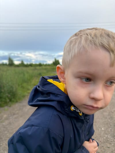 The media file is a close-up, rotated photo of a young child with blonde hair. The child is wearing a dark blue jacket with yellow accents on the zipper pulls and collar. The child's face is turned towards the camera, with a slight tilt to their head. They have blue eyes and fair skin. The background is blurred, showing a grassy field and a cloudy sky, suggesting an outdoor setting, possibly in the countryside. The lighting suggests it might be daytime, possibly late afternoon or early evening, given the soft light. The child's expression is neutral or slightly curious. There is no visible text in the image. The provided location context is "Чёрная, Russia".