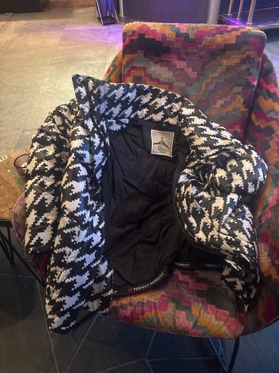 The image displays a black and white houndstooth puffer jacket resting on a colorful armchair. The jacket is open, revealing its black interior lining. A white rectangular label is sewn into the upper back of the jacket's lining, featuring a star emblem and the brand name "PERFECT MOMENT" written in black text. Additionally, a black fabric trim or band, visible along the inside edge of the jacket, repeatedly displays the text "PERFECT MOMENT" with a small red star between the words.
The armchair on which the jacket is placed is upholstered with a vibrant, multi-colored fabric featuring a zigzag or chevron pattern in shades of purple, dark red, orange, brown, and green. The chair appears to have sturdy armrests, although they are mostly obscured by the jacket.
The background shows a dark, possibly tiled or stone floor, with a slight sheen or reflection. A dark metal furniture frame or leg is partially visible on the right side of the image. The overall setting appears to be indoors. The image itself is rotated approximately 90 degrees counter-clockwise.