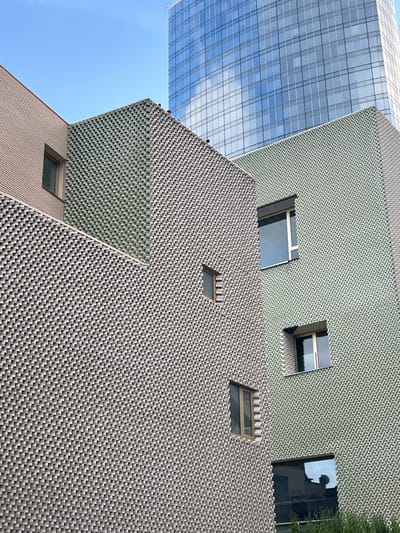 This image shows a portion of the exterior of the AGInsurance headquarters in Saint-Josse-ten-Noode - Sint-Joost-ten-Node, Belgium. The building features a distinctive facade composed of intricately patterned bricks in shades of beige and green. Several windows are visible, some rectangular and others with more angular frames. A large, modern skyscraper with a glass facade is partially visible in the background, reflecting the blue sky and clouds. The foreground shows the textured brickwork of the lower buildings. There are no people or identifiable figures in the image.