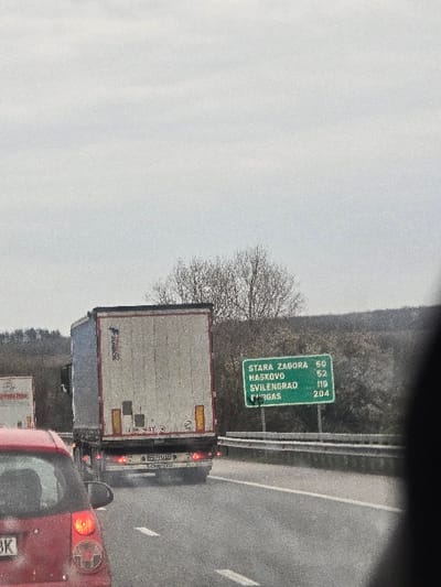 The scene depicts a multi-lane highway in the vicinity of Plodovitovo, Bulgaria, under an overcast, light gray sky. In the immediate foreground on the left, the rear section of a red passenger car is visible, featuring an illuminated left taillight and a partial license plate ending in "BK". Directly ahead, occupying the central midground, a white semi-trailer truck travels forward. The truck's trailer displays faint branding, possibly "SCHMITZ," along with wear marks and illuminated rear lights. Further in the background on the left, a portion of a second white semi-trailer truck is visible. To the right of the central truck, a prominent green highway sign with white text is mounted, indicating destinations and distances in kilometers: "STARA ZAGORA 50", "HASKOVO 52", "SVILENGRAD 119", and "BURGAS 204". The highway features asphalt paving with distinct white lane markings. A metallic guardrail runs along the right edge of the road, separating it from an upward-sloping, vegetated embankment. The background beyond the road features undulating hills covered with deciduous trees, many appearing bare, under the uniform cloud cover. No persons are visible within the frame.