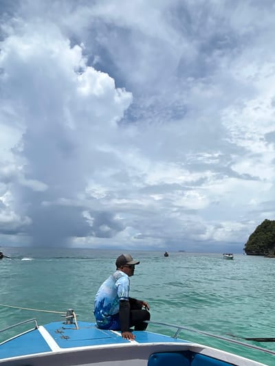 A man wearing a cap and a blue t-shirt with a mountain graphic is sitting on the bow of a blue and white boat. He is facing to the right, looking out at the ocean. The water is turquoise and there are several other boats in the distance. The sky is cloudy with some patches of blue. In the background, there is a green, rocky island. The scene appears to be in the waters near Ko, Thailand.