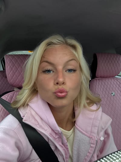 The scene depicts a young blonde female seated within the interior of a vehicle, located in the city of Iver, United Kingdom. She possesses blue eyes and long blonde hair, styled to frame her face. Her expression is characterized by pursed lips, forming a "kiss" face, as she looks directly towards the camera. She is attired in a light pink hooded jacket with a zippered front, worn over a cream or light yellow ribbed top. A thin, delicate necklace is visible around her neck. A black seatbelt is fastened diagonally across her torso and horizontally across her lap. The vehicle's interior features custom pink, diamond-quilted seat covers on all visible seats, including the front passenger seat to her right and the rear seats in the background. A small, metallic, crown-shaped embellishment is affixed to the headrest area of the front passenger seat cover. The car's headliner is dark grey. The lighting appears even, illuminating the subject and interior.