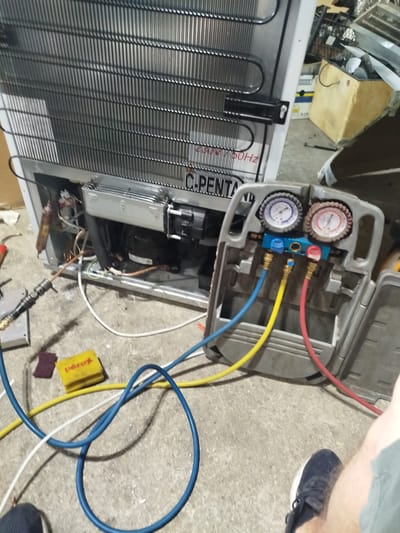 The image shows a close-up view of the back of a refrigerator, with its condenser coils prominently displayed. A manifold gauge set with blue, yellow, and red hoses is connected to the refrigerator's compressor and refrigerant lines. The scene appears to be in a workshop or garage, with concrete flooring and various tools and equipment scattered around. The ambient lighting suggests it might be daytime, but the specific location within the city of Prijedor, Bosnia and Herzegovina, cannot be determined from the visual information provided. A person's leg and foot are visible in the lower right corner, wearing a dark blue athletic shoe, indicating human presence and likely engagement with the equipment. The refrigerator itself is partially visible, with its branding including "Penta".