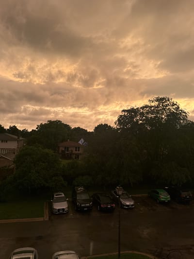The image captures a residential street in Glendale Heights, United States of America, under a dramatic, cloudy sky with hues of orange and yellow, suggesting either sunrise or sunset. The foreground displays a wet asphalt surface, indicating recent rain, with several parked cars. To the left, partially visible, are the back ends of two cars, one light silver and another dark. Further into the scene, a light silver sedan, possibly an Audi, is parked in a designated spot. Next to it is a dark SUV. Beyond the parking area, a line of lush green trees forms a dense backdrop, partially obscuring residential buildings. One building, with a brown roof and a light-colored facade, is visible in the mid-ground. The overall atmosphere is somber, amplified by the overcast sky and the dark, wet surroundings.
