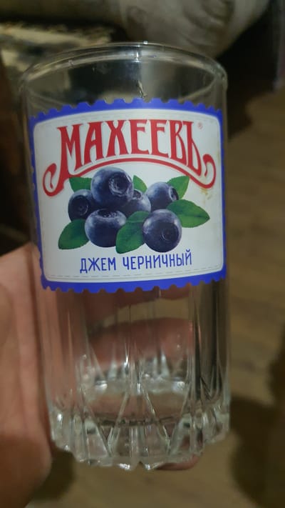 A glass of clear liquid sits on a wooden surface. A label on the glass reads "МАХЕЕВЪ" in red lettering above an illustration of blueberries. Below the illustration, in blue lettering, it says "ДЖЕМ ЧЕРНИЧНЫЙ" which translates to "blueberry jam." The glass is transparent with a textured pattern at the bottom. The background is out of focus but appears to be a room. There is no indication of the city of Karankul, Uzbekistan.