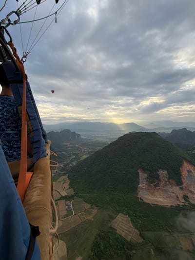 An eye-level, wide-angle shot captures a hot air balloon ride over a rural landscape at dawn. The foreground is dominated by the interior of a hot air balloon basket, showing the back of a person in a blue shirt with a geometric pattern and a portion of the balloon's rigging. Orange straps and brown material of the basket are visible. In the mid-ground, two other hot air balloons are visible in the hazy sky, one red and the other dark. The background reveals a valley with rolling green hills, agricultural fields, and distant mountains shrouded in mist. Sunbeams break through the clouds, illuminating the scene. The location is identified as Hong Ngern Village, Laos.