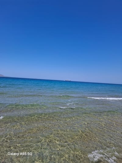 The image captures a serene coastal scene under a clear, bright blue sky in the Gouves Municipal Unit, Greece. The foreground features transparent, shallow turquoise-green sea water, revealing a light-colored, textured seabed with visible rocks and sand. Gentle waves ripple across the surface, some breaking into white foam at the bottom right. The midground shows the deeper, darker blue expanse of the Aegean Sea, with a prominent white and blue passenger ship or ferry sailing horizontally from left to right in the distance, positioned slightly above the horizon line. Further to the left, a distant, light brown mountainous landmass or island is faintly visible on the horizon. The top half of the image is dominated by an uninterrupted, vivid azure sky. There are no people present in the scene. A white watermark "Galaxy A55 5G" appears in the bottom left corner of the image.