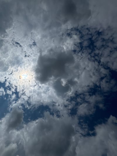 The image is a low-angle, full-frame shot of a cloudy sky. The clouds are varied in density and color, ranging from dark, ominous gray masses to lighter, more wispy formations. The sun is partially visible in the upper left quadrant, peeking through a break in the clouds, casting a bright, diffused light with some iridescent colors. The sky appears to be in a state of transition, possibly indicating impending weather changes. There are no people, buildings, or any other discernible objects or landmarks visible in the frame, solely the natural atmospheric conditions. The location in the city of Tashkino, Russia, is not visually discernible from the image itself.