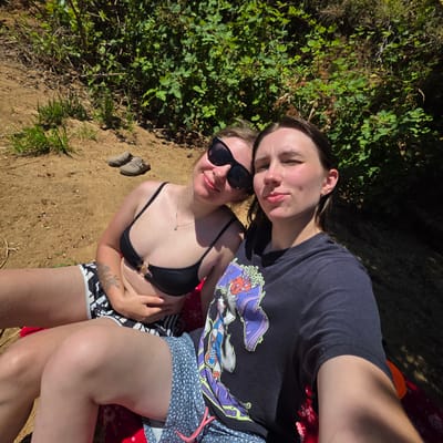 In Cherry Grove, Oregon, two individuals are captured in a selfie taken outdoors. They are positioned on the ground, with lush green foliage in the background and a sandy, earthy terrain surrounding them. The person on the left is wearing a black bikini top and patterned bottoms, with sunglasses resting on her head. The person on the right is wearing a dark t-shirt with a graphic print. Both appear relaxed and are enjoying the natural environment. A pair of casual shoes are visible in the background, suggesting they may have been walking or hiking prior to taking this photo. The lighting indicates it is daytime, with sunlight filtering through the trees, creating dappled shadows.