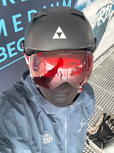 A person is depicted from the chest up in a close-up, selfie-style shot within a ski resort environment. The individual wears a black ski helmet with a white Fischer logo (three interlocking triangles) on the front, red-tinted Oakley ski goggles, and a black balaclava or neck gaiter. A blue-grey hooded ski jacket, featuring a white circular Gastein logo on the left chest and a faint Arc'teryx logo on the right chest, completes their attire. To the lower right, a pair of black and white ski gloves rests on a textured, possibly metallic, patterned floor. The goggles reflect an outdoor scene with snow, multiple individuals (some appearing to be skiers), parts of a building or lift station, and the photographer's hand holding the camera. The background behind the person includes a dark blue wall or banner with partial light blue text ("MER BEC") on the left, and a banner depicting a snowboard/ski graphic against a blue and white sky on the right. The "Gastein" logo on the jacket indicates the location is within the Gastein Valley ski region, specifically in or around Bad Gastein, Austria.