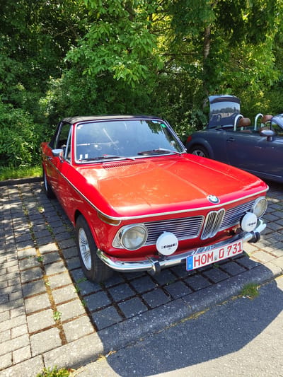 A bright red convertible BMW is parked on a cobblestone surface in front of a dense line of green trees. The car has a black soft top and chrome accents, including its front grille, headlights, and bumper. Two large, round fog lights with "CIBIE LED" branding are mounted on the front bumper. The car's license plate reads "HOM D 731". Behind the red BMW, a dark blue convertible is partially visible, with its hood open, revealing brown leather seats. The scene appears to be a sunny day with strong shadows cast on the ground. The location is the city of Pirmasens, Germany.