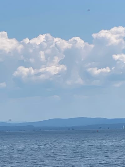 The image captures a scenic view of a body of water next to a coastline, with a bright blue sky filled with fluffy white clouds overhead. The water has a textured appearance, suggesting gentle waves or ripples. In the distance, faint landmasses are visible, appearing as soft blue layers that recede into the horizon, hinting at a hazy atmosphere or the curvature of the Earth.
On the distant shoreline, small structures or trees are subtly discernible. A tiny red object, possibly a marker or a buoy, is visible on the water near the coast. There are also what appear to be small white specks in the distance, which could be sailboats or distant objects. One such speck, a small dark square, is in the upper left portion of the sky. The overall scene suggests a peaceful day, likely during daylight hours given the bright sky and clouds. The location is described as the Town of Peru, United States of America, though no specific landmarks within the town are identifiable from this image. The weather appears to be clear and pleasant, conducive to outdoor activities.