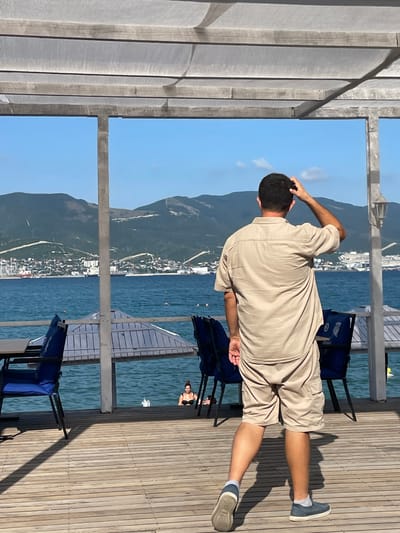 A man stands on a wooden pier, his back to the camera, with his hand on his head. He is wearing a light-colored short-sleeved shirt and beige shorts. His left leg is extended, and his right foot is slightly bent. The pier has a wooden plank floor with visible grain and shadows. To the left of the man, a gray, slatted structure provides shade.

In the background, a town is visible nestled at the base of a large, green, mountainous landscape. The town appears to have buildings with white walls and red roofs. Beyond the town, the blue sea stretches out, with gentle waves. The sky is a clear, bright blue with a few wispy white clouds.

Several blue chairs with dark frames are positioned on the pier. One chair is visible behind the man, and another is further to the right. In the water, a person in a black bikini can be seen, looking towards the camera. The overall scene suggests a sunny day, likely in the late morning or early afternoon, given the strong shadows. The location appears to be a coastal area with a town and mountainous backdrop, consistent with the provided location context of Varnavinskoe, Russia.