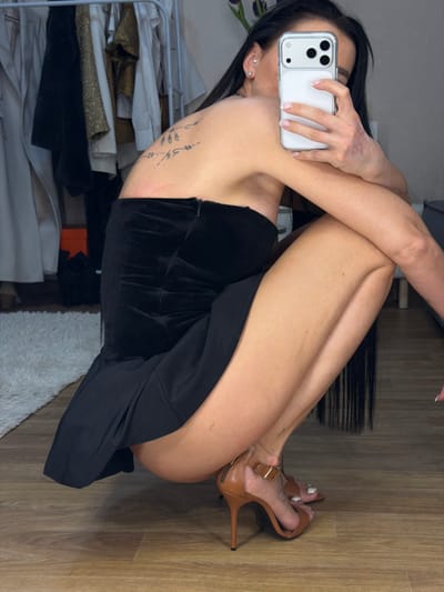 The image depicts a female subject capturing a mirror selfie indoors, in a crouching position on a light-toned wooden floor. She is wearing a strapless black garment, comprising a velvet-textured upper section and a flowing skirt, paired with brown high-heeled sandals featuring ankle straps. Her face is obscured by a white-cased smartphone, equipped with a multi-lens camera module, which she holds in her left hand. The subject has long dark hair, visible small stud earrings in her right ear, and a linear tattoo consisting of organic, leaf-like designs on her upper back/shoulder blade. Her fingernails and visible toenails are painted with a light-colored polish.
In the background on the left side of the frame, a metal clothing rack holds various garments, including light-colored coats and a shimmering gold item. A portion of a white shag rug is visible on the floor adjacent to the clothing rack. Above the rack, a dark floral arrangement is discernible in a vase. On the far right, a dark grey upholstered furniture piece is partially visible. The walls in the room are light in color. The specific location within the city of совхоза "Воронежский", Russia, cannot be determined from the visual information provided in the image.