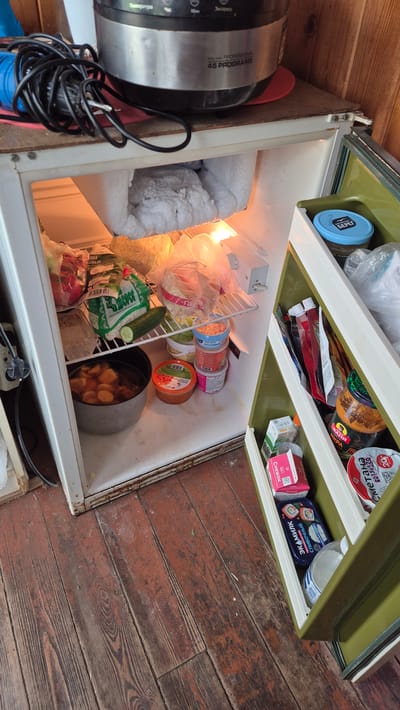 This is a medium shot, eye-level, exterior photograph of an open, old, white, two-door refrigerator. The refrigerator is situated on a wooden floor, likely in a rustic or outdoor setting. The upper compartment appears to be a freezer, with significant frost accumulation on the back wall and items below. The main compartment of the refrigerator is filled with various food items. On the top shelf, there are plastic bags containing produce, including what appears to be cabbage and radishes. A cucumber is also visible on this shelf. Below this, there is a large pot containing food, possibly stewed potatoes, and several small containers of what look like spreads or dips. The refrigerator door itself has multiple shelves, stocked with jars, packets, and containers of food, including what appear to be yogurt or sour cream containers, and a jar with a red label that might be jam or sauce. The overall impression is of a functional, albeit somewhat aged, refrigerator containing a variety of food staples. The setting suggests a practical storage solution in an environment where a modern kitchen may not be available. The wooden floor boards are worn and appear to be aged, contributing to the rustic aesthetic. The presence of a multi-cooker on top of the refrigerator indicates it may be placed in a temporary or auxiliary kitchen area. The specific location of Машезеро, Russia, suggests a rural or possibly dacha (summer house) setting.